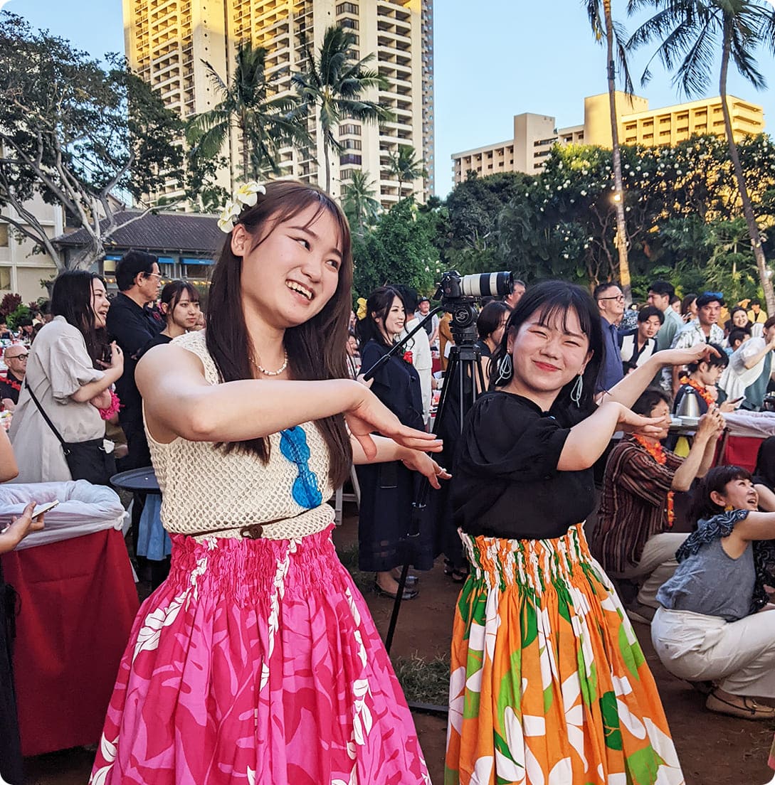 屋外でダンスを楽しむ2人の女性社員。カラフルなスカートを履いて、笑顔でハワイアンのようなポーズをとっている。