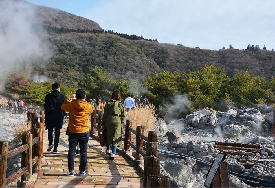 温泉地の小道を歩く観光客たち。周囲には蒸気が立ち上っている。